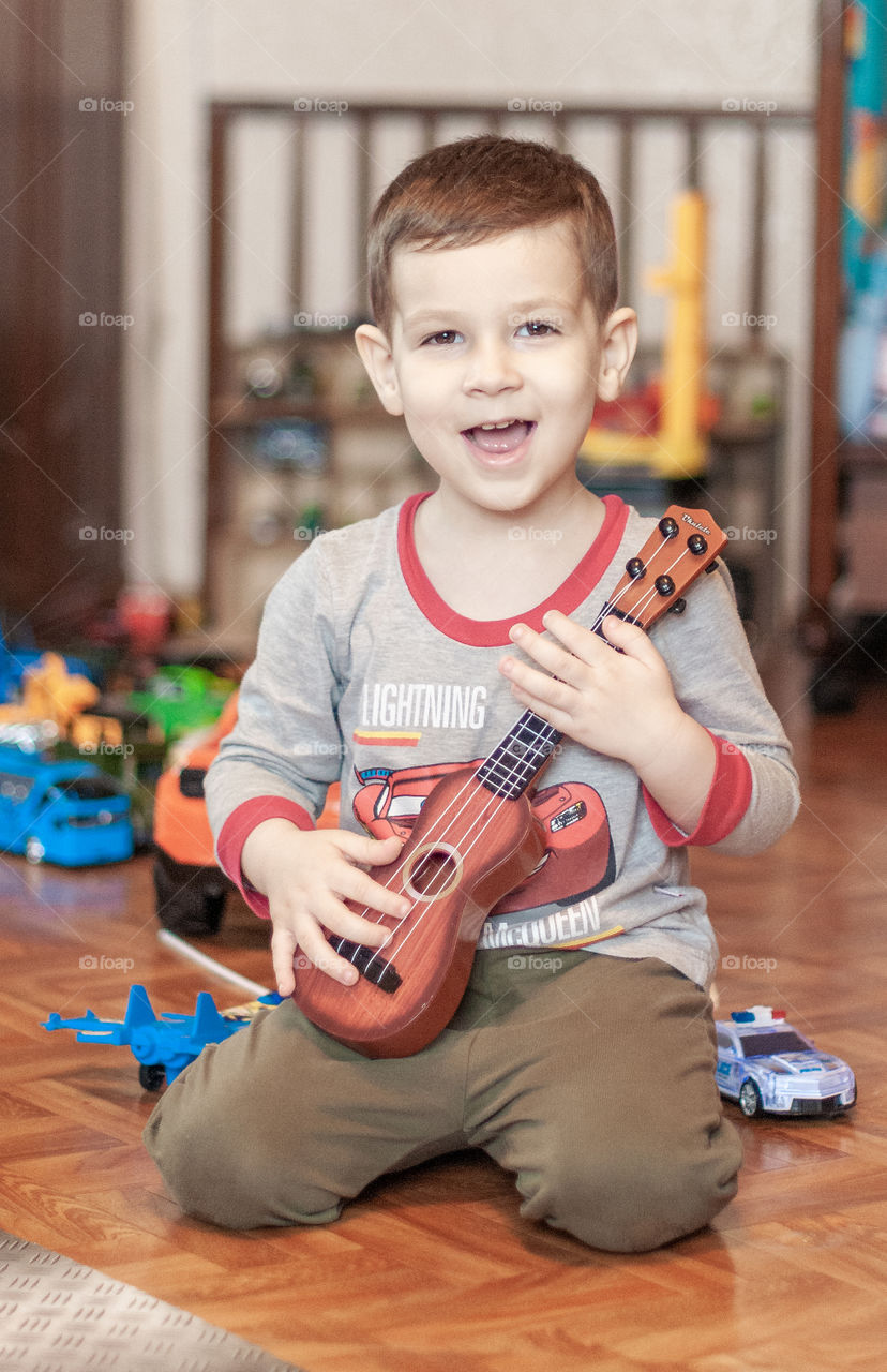 Toddler boy sitting at the floor at home and playing the little guitar 