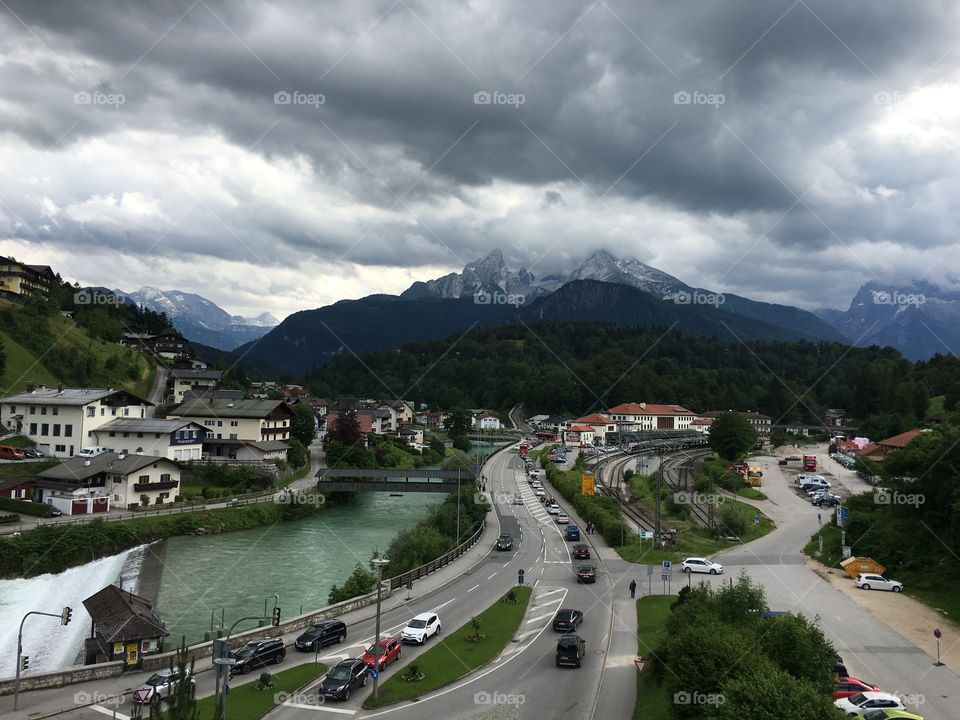 Large dark clouds loom over a small town in the German Alps, throwing shadows on a clear turquoise river, train station and road. 