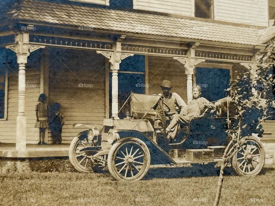 Man and son in old car in front of old house with other children in shadows in porch