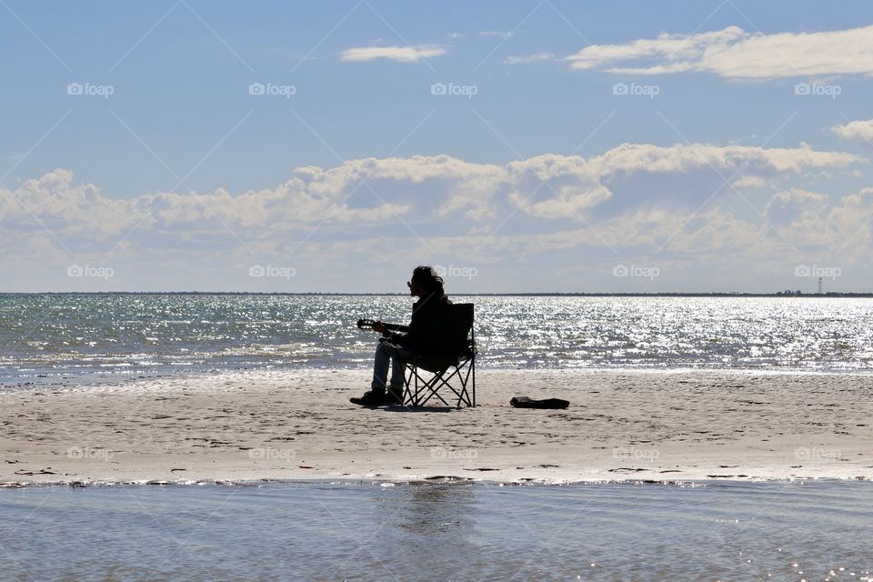 Silhouette of male musician guitarist singer sitting in chair surrounded by glistening ocean performing solo 