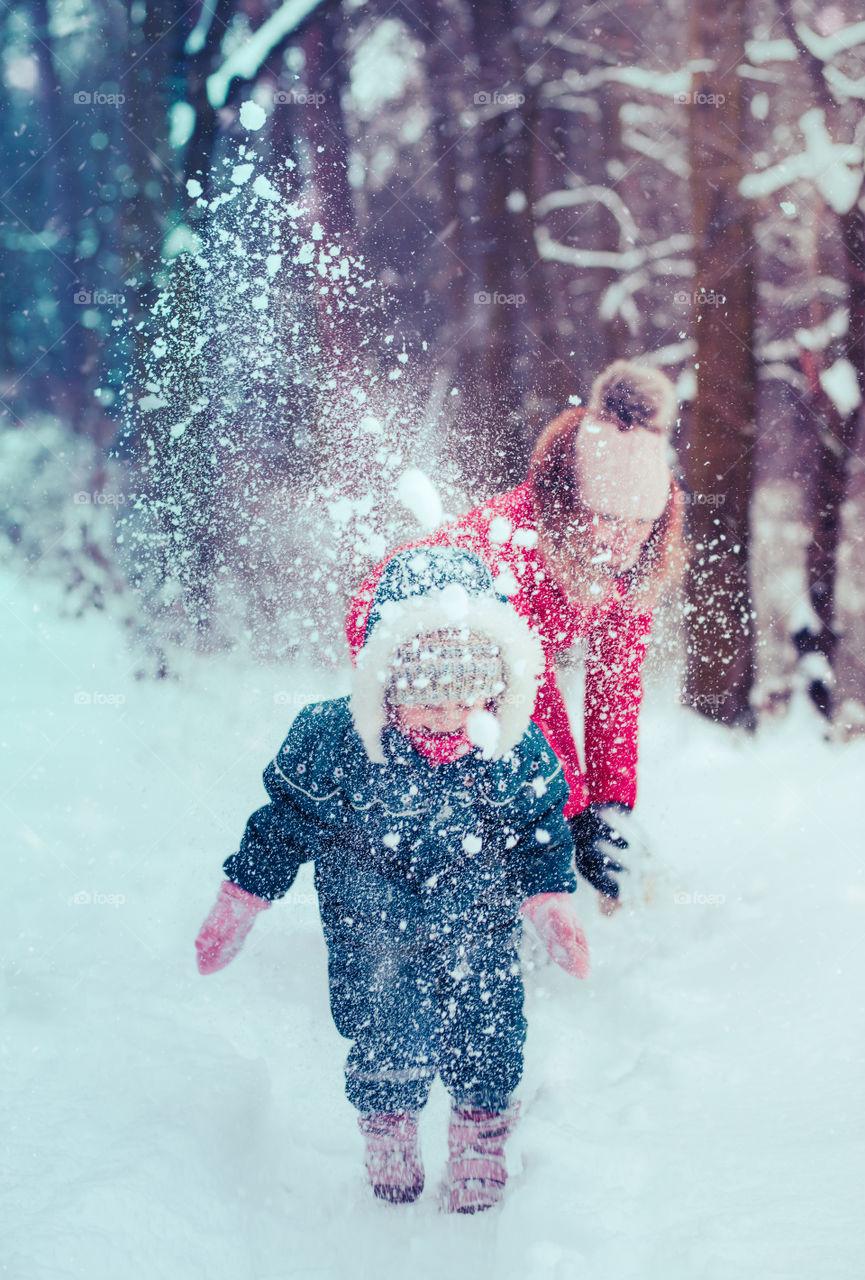 Mother and her little daughter are spending time together walking outdoors in forest in winter while snow falling. Woman is pulling sled, a few years old girl is walking through the deep snow, enjoying wintertime