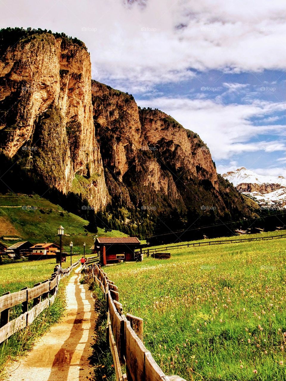 Landscape of the Dolomite mountains looking down a valley with a winding fenced path, large craggy  golden hued mountains and snow capped mountains in the distance