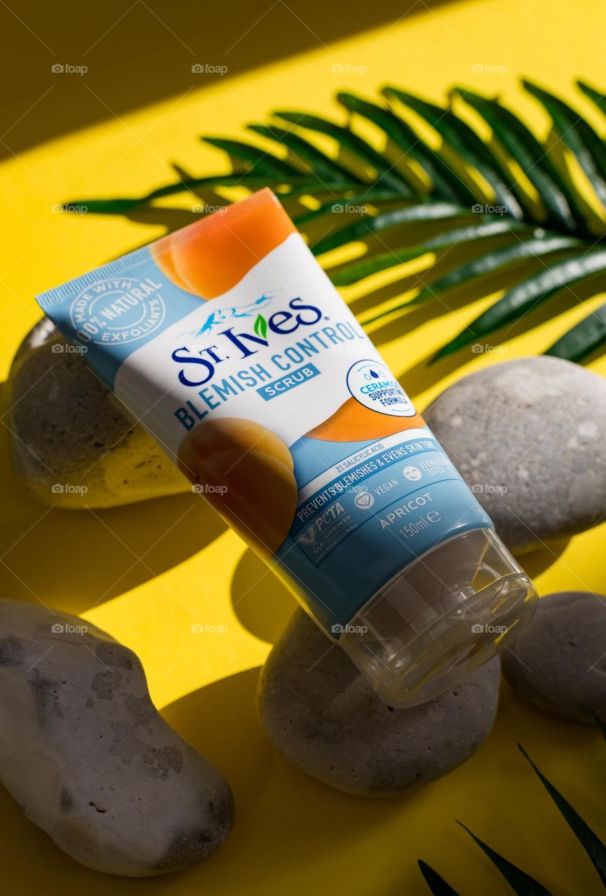 One tube of natural apricot scoab brand st. Ives lies on gray stones with palm branches on a yellow background with shadows and light from the sun, close-up side view. The concept of female cosmetics, body care, beauty