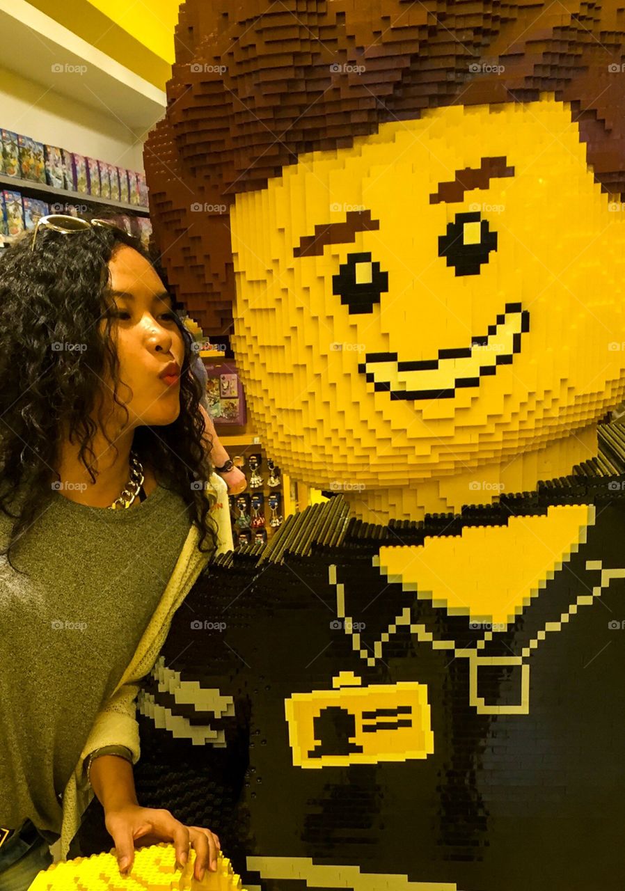 Girl with curly hair wearing a gray T-shirt, have a sunglasses on her hair is kissing Mr. Lego.  He is smiling because of happiness