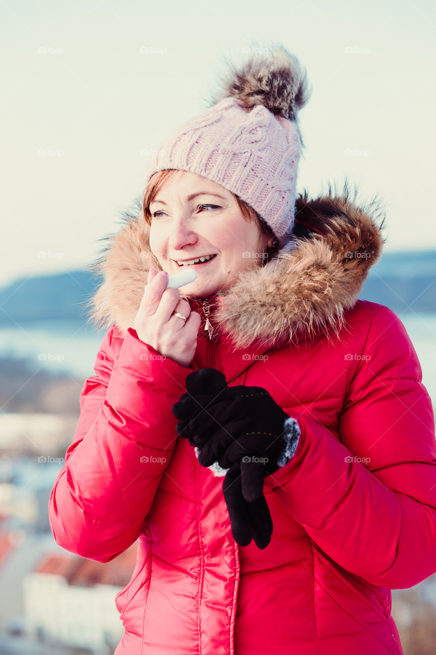 Woman applying lip balsam while walk on a wintery day. Wearing red coat and cap. Town in the background