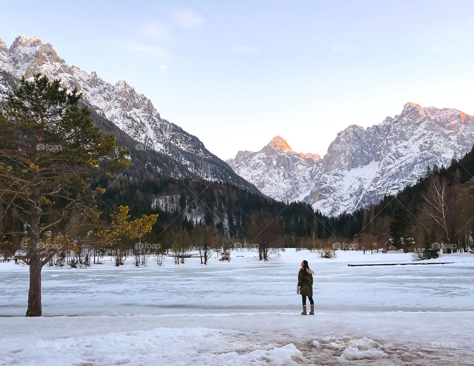 Winter is here, icy lake with incredible view of mountains covered with golden sunlight, female person standing by lakeside, enjoying the view