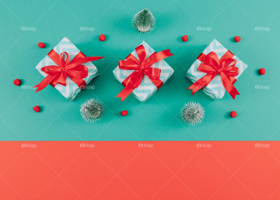 Three gift boxes with red ribbons, decorative Christmas trees and fluffy snow on a red-green background with copy space from below, flat lay close-up. Holiday gifts concept.