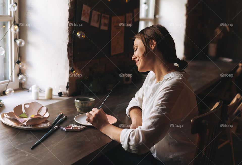 Young attractive woman in white shirt ceramic artist decorating clay plate with tool at table in pottery workshop. Handmade work student, freelance small business