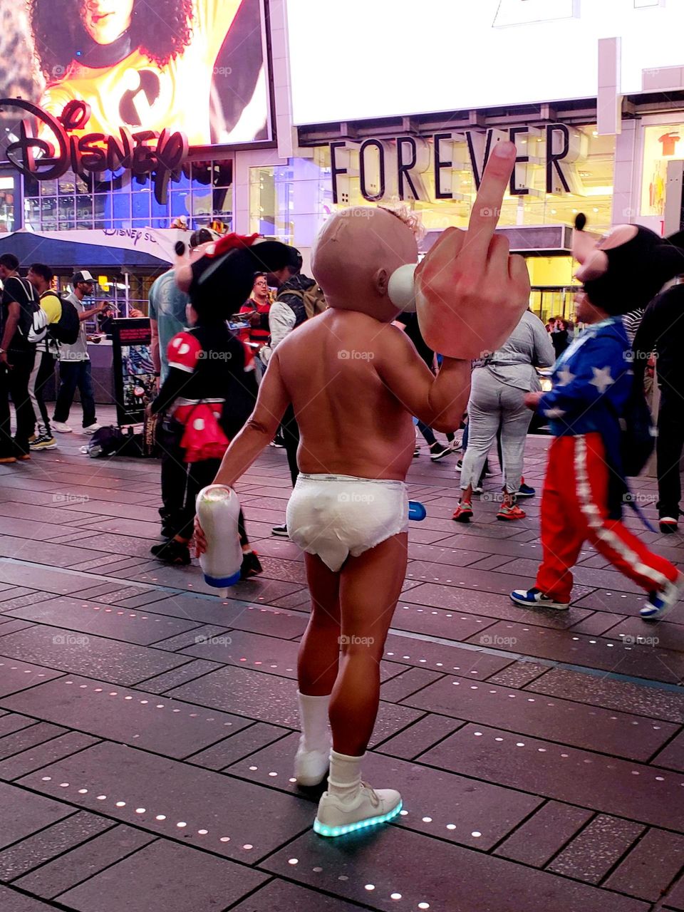 Times Square New York City. An adult is dressed in character as a baby, wearing only a diaper, shoes, and socks. on his hand is a large foam glove showing off the middle finger.