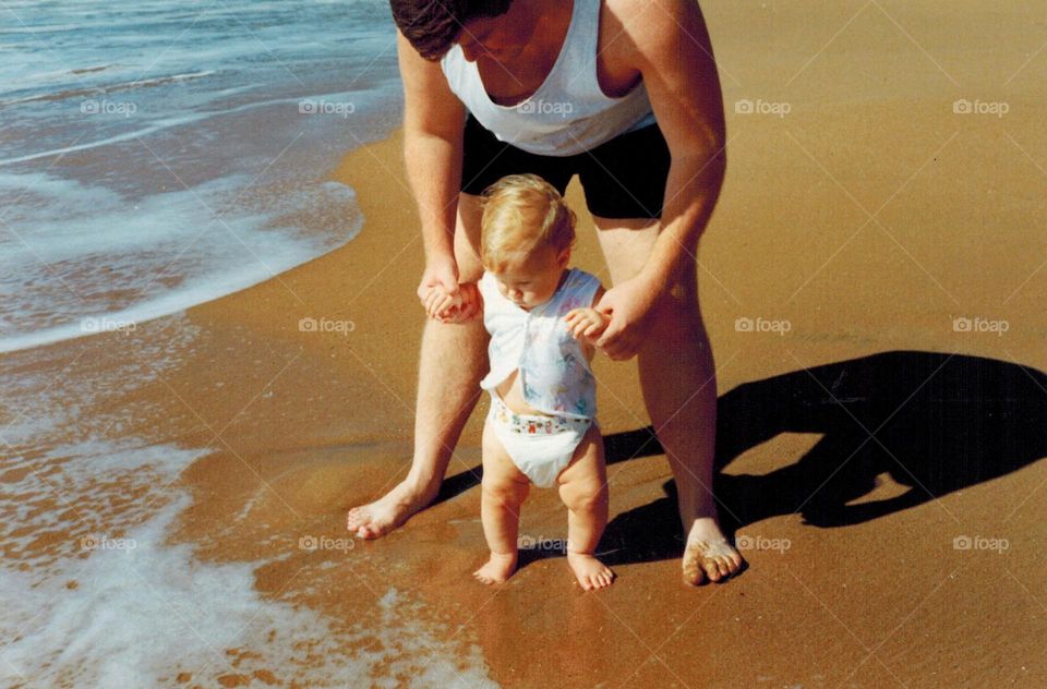 Father leads baby daughter by the hand on first few steps to the shore