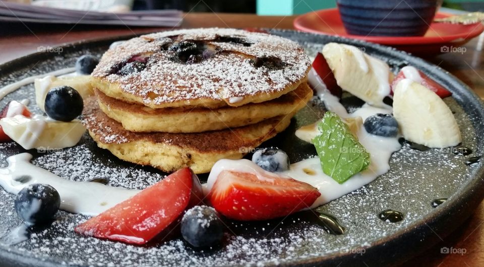 Pancakes and fresh fruit on black plate dusted with icing sugar.