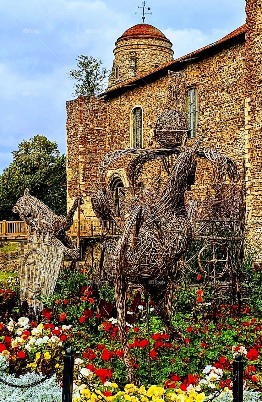 Large wicker figure of a Knight on horseback in a bed of bright red and yellow flowers in front of Colchester castle