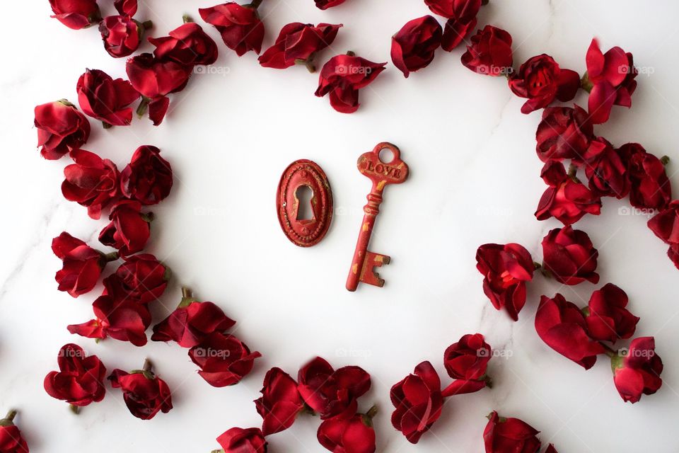 Red, metal vintage key and keyhole with red, silk rose buds in a heart-shaped arrangement on white marble surface