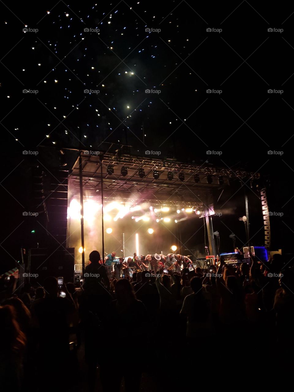 Night-time concert coming to an end as the crowd cheers in appreciation and fireworks begin in the background.