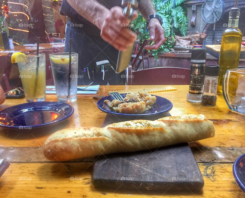 A loaf of Italian bread sitting on a rustic wooden table in a restaurant while the server prepares a garlic dip in the background.