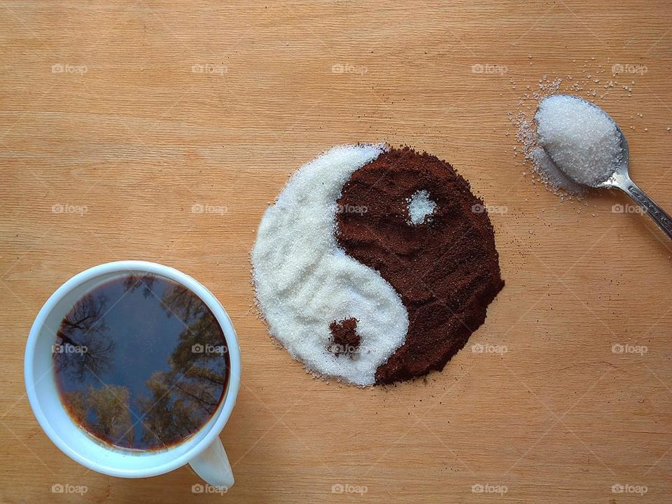 On a wooden background there is a white cup with black coffee in which the tops of the trees are reflected, a yin-yang symbol made of white sugar and brown ground coffee, and a spoon with white sugar. Philosophy