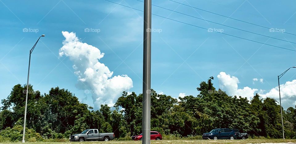 Commuting: vehicles behind the metal grey pole in front of the green trees and seeing a blue sky with clouds and cumuli nimbus, driving in front of each other in a pickup truck, SUV and car, grey, red and blue.