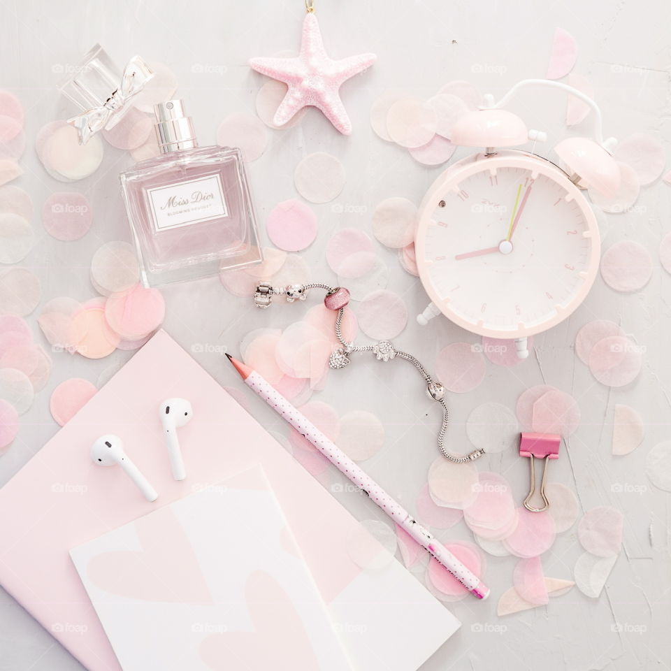 Flatlay items: alarm clock pink color, perfume, notebook, pencil, wireless headphones, confetti, bracelet, star lying on gray concrete background 