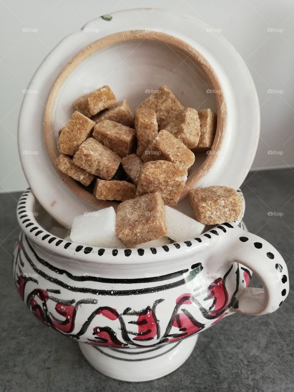 Multicoloured and patterned sugar container with handle and top and striped leg is full of pieces of white and brown sugar