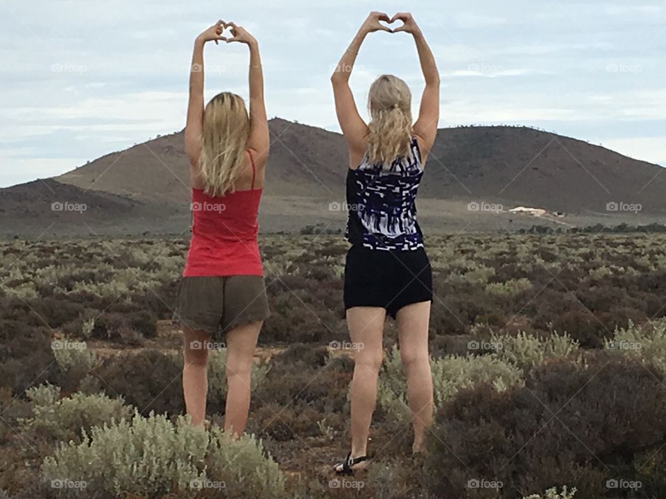 Freedom in wide open spaces, two teenage blonde girls with arms upraised hands in heart position facing mountain In Remote Australian outback of south Australia 