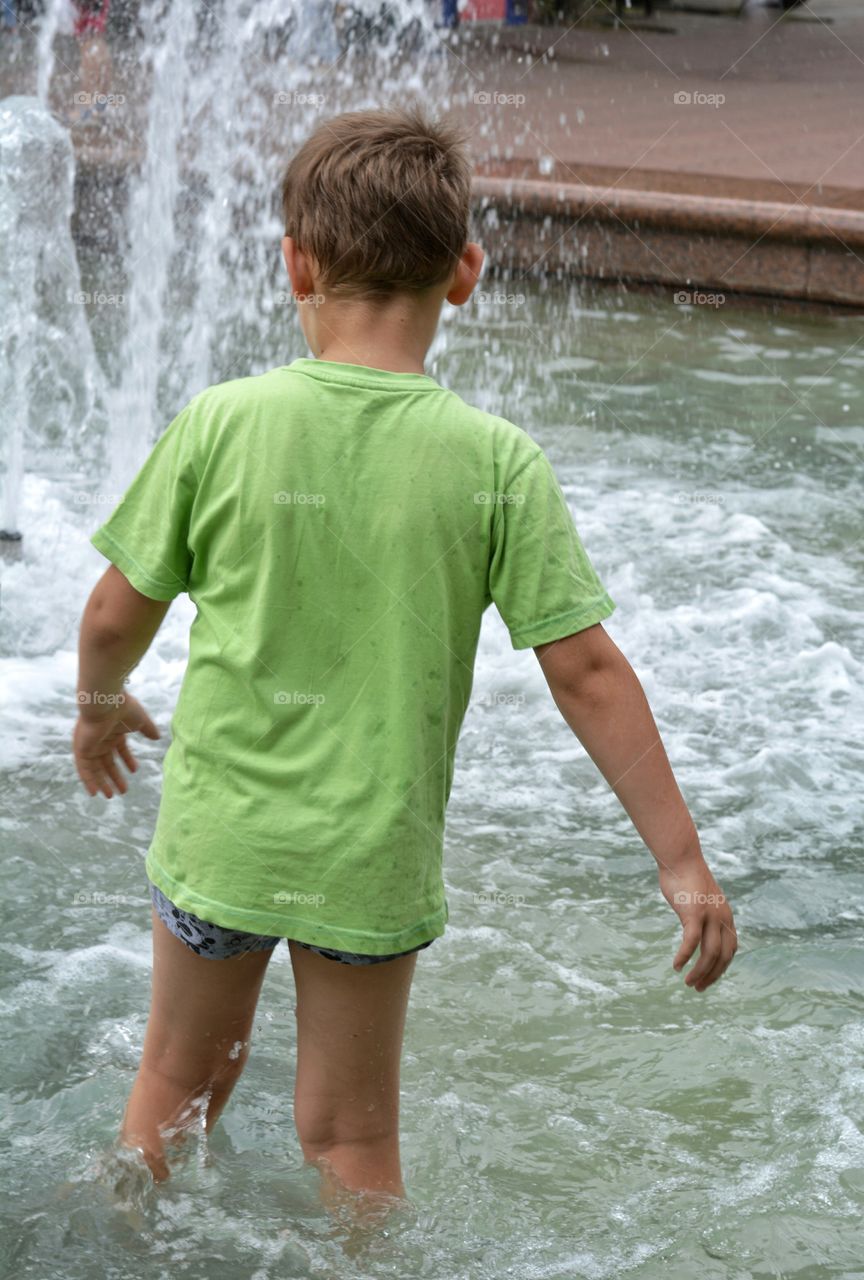 child boy in the water splash fountain, summer heat, city street view