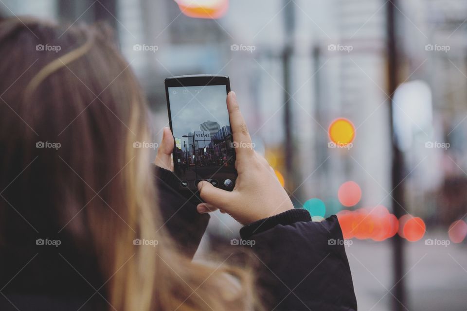 A girl on her phone taking a photo of the scene, focused on her hands and the phone photo with lights in the background