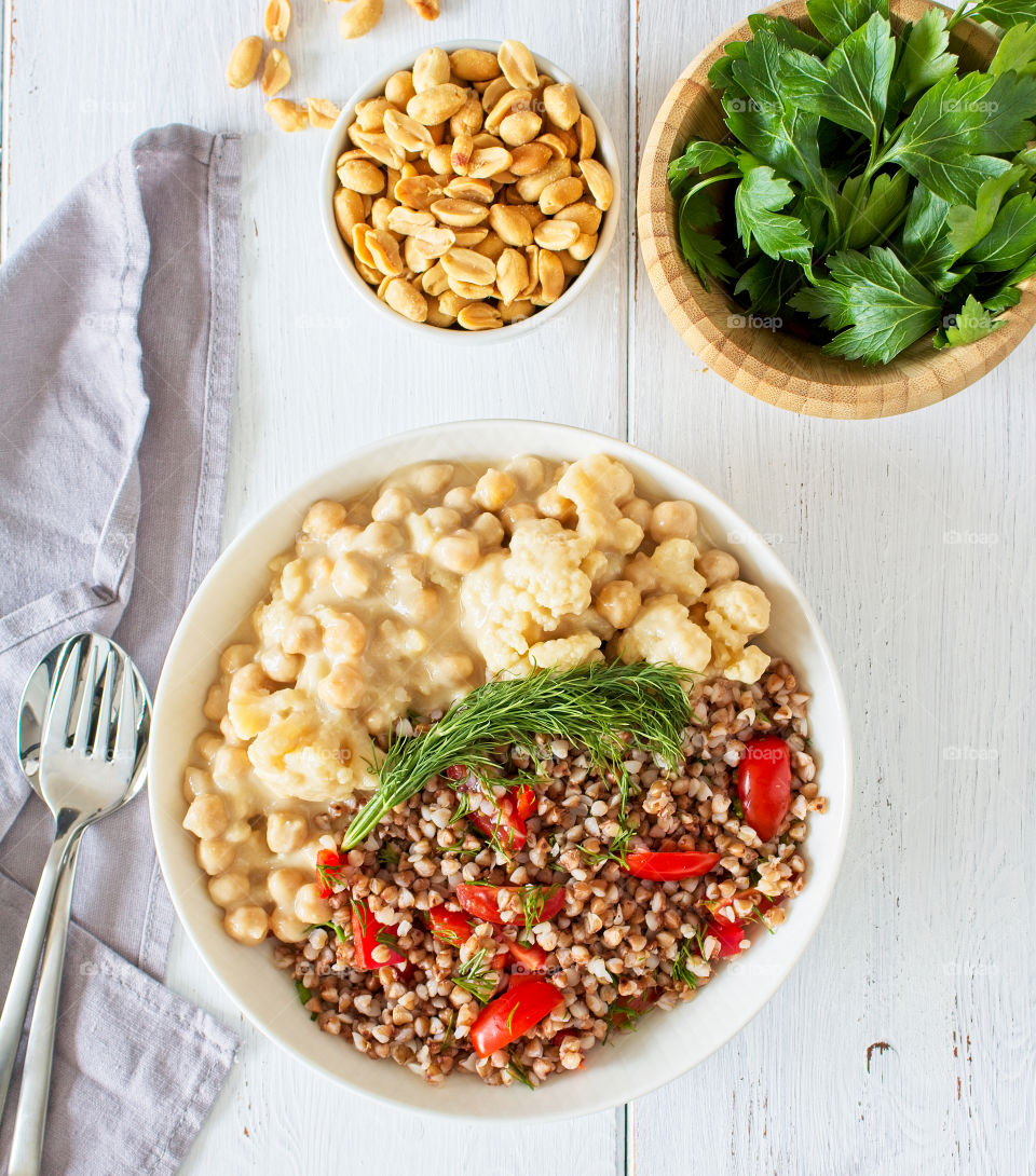 Cauliflower with chickpeas in peanut sauce in a bowl with buckwheat and red peppers, decorated with greens. Some greens and peanuts are next to the bowl. Vegan healthy lunch