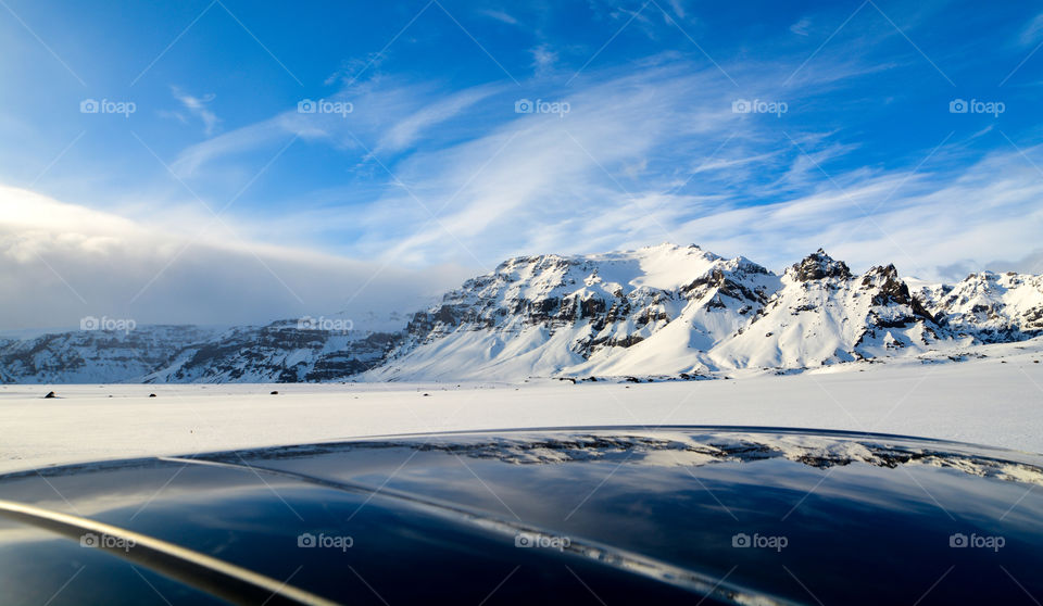 Snowy mountain range in Iceland under blue sky with its reflection on a car hood