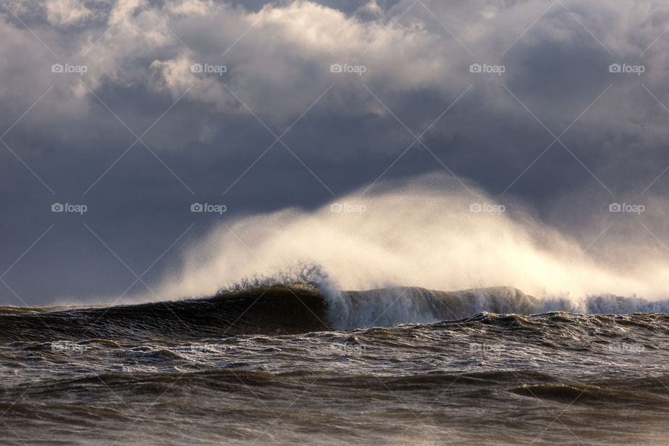 Large wave breaking with white spray being lit up by sunlight against a stormy cloudy sky.