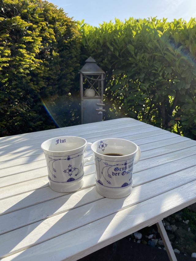 Sunrise behind the hedge, two cups of fresh coffee on the white wooden table in the morning at the Baltic Sea . A candle foarms the axis of the image 