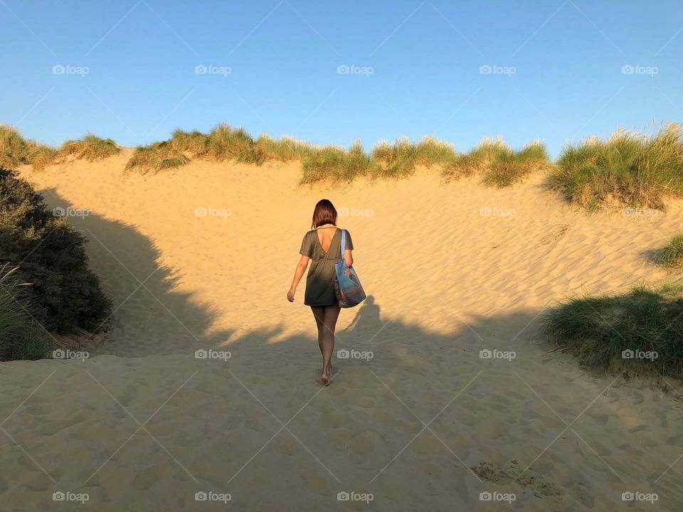 A girl in a tunic and with a beach bag walks along a sand dune. Sunny summer day on the beach.