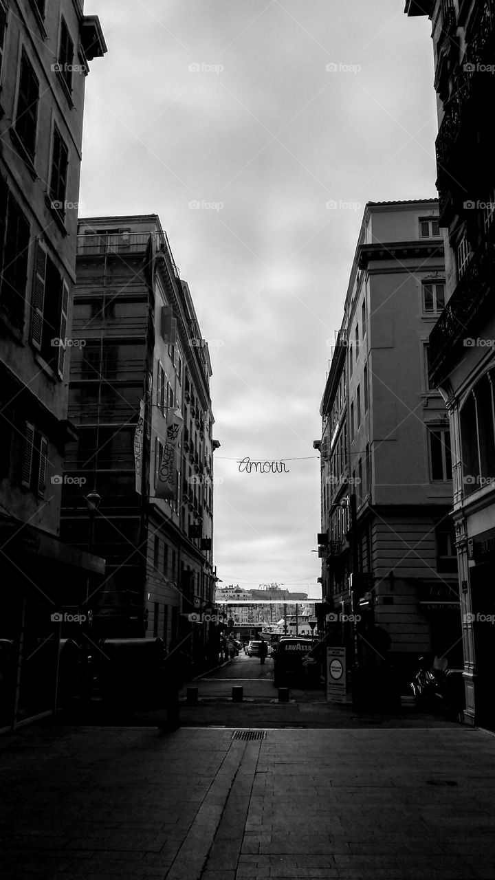side of buildings separated by a cobblestone street, in the middle of them and at the top hangs a sign with the
French word "love". In the background you can see the city old port and a cloudy sky.