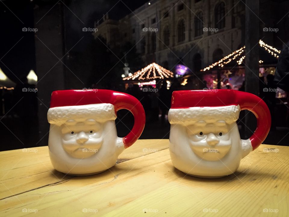 Two mugs shaped like Santa Claus on wooden table with Christmas lights in the background 
