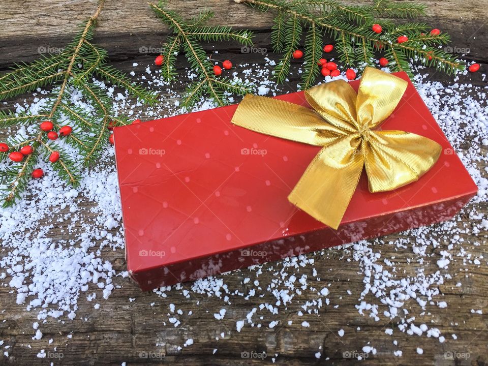 Gift wrapped in red paper with a golden ribbon on wooden table powdered with snow and pine cone tree branches 