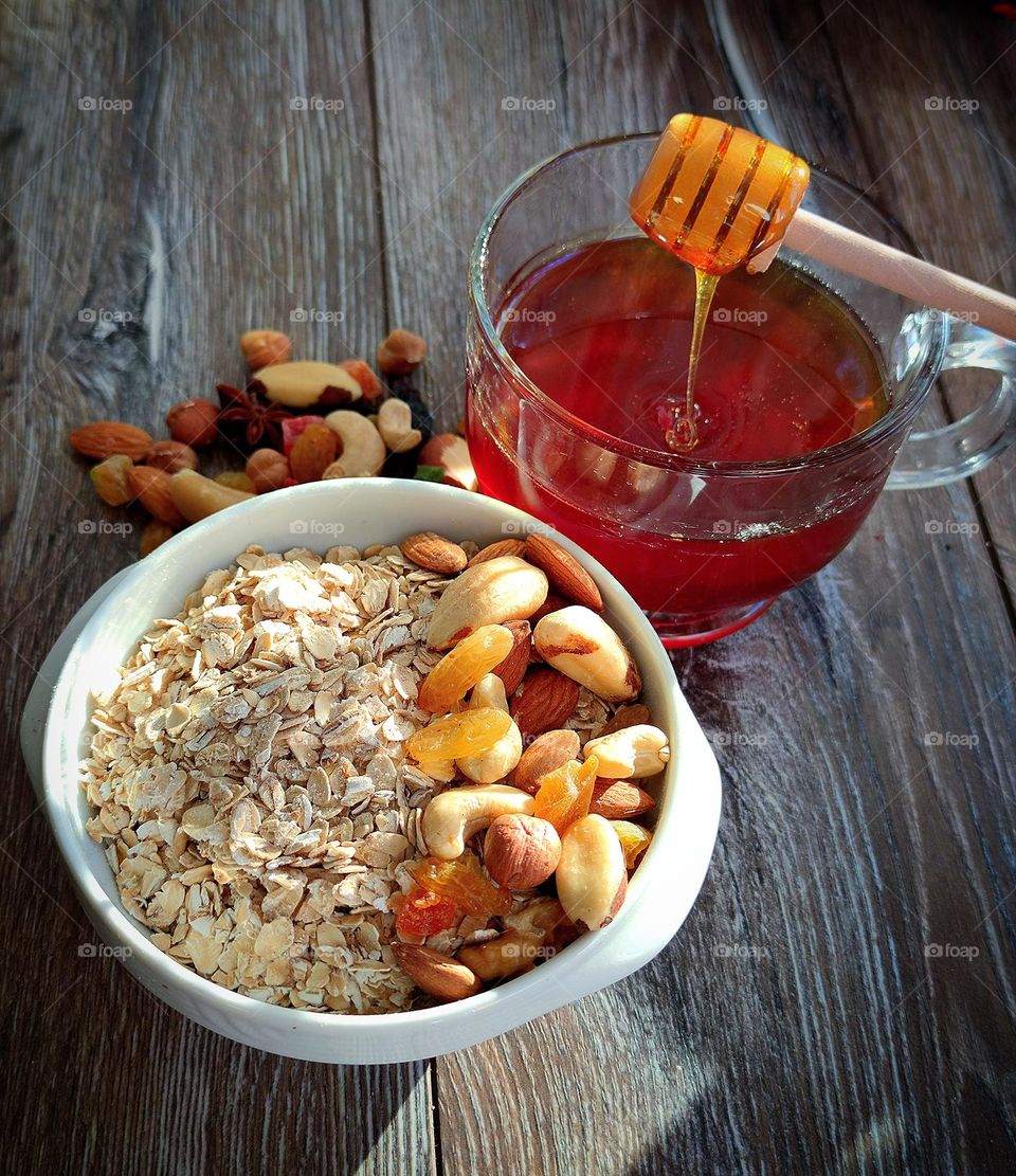 Food.  On a wooden background is a white plate with oatmeal and a nut mix with candied fruits.  Nearby is a mug with honey and a wooden twisted spoon from which honey flows.  Behind the plate lies a slide of nut mixture