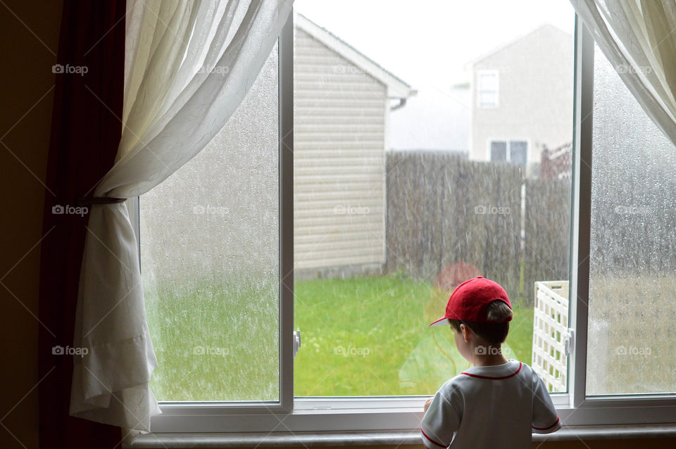 Young child toddler boy wearing a baseball cap and watching the rain out of a window of a home