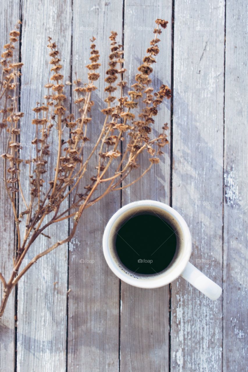 Flat lay of coffee in a white mug next to a dried brown plant on a weathered wooden surface