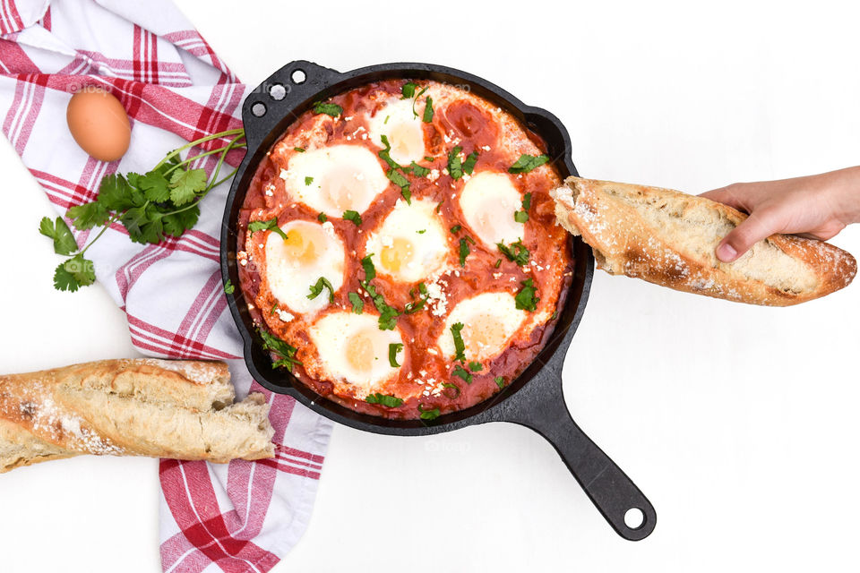 Flat lay of a shakshuka dish prepared in a cast iron skillet with a person's hand holding a baguette