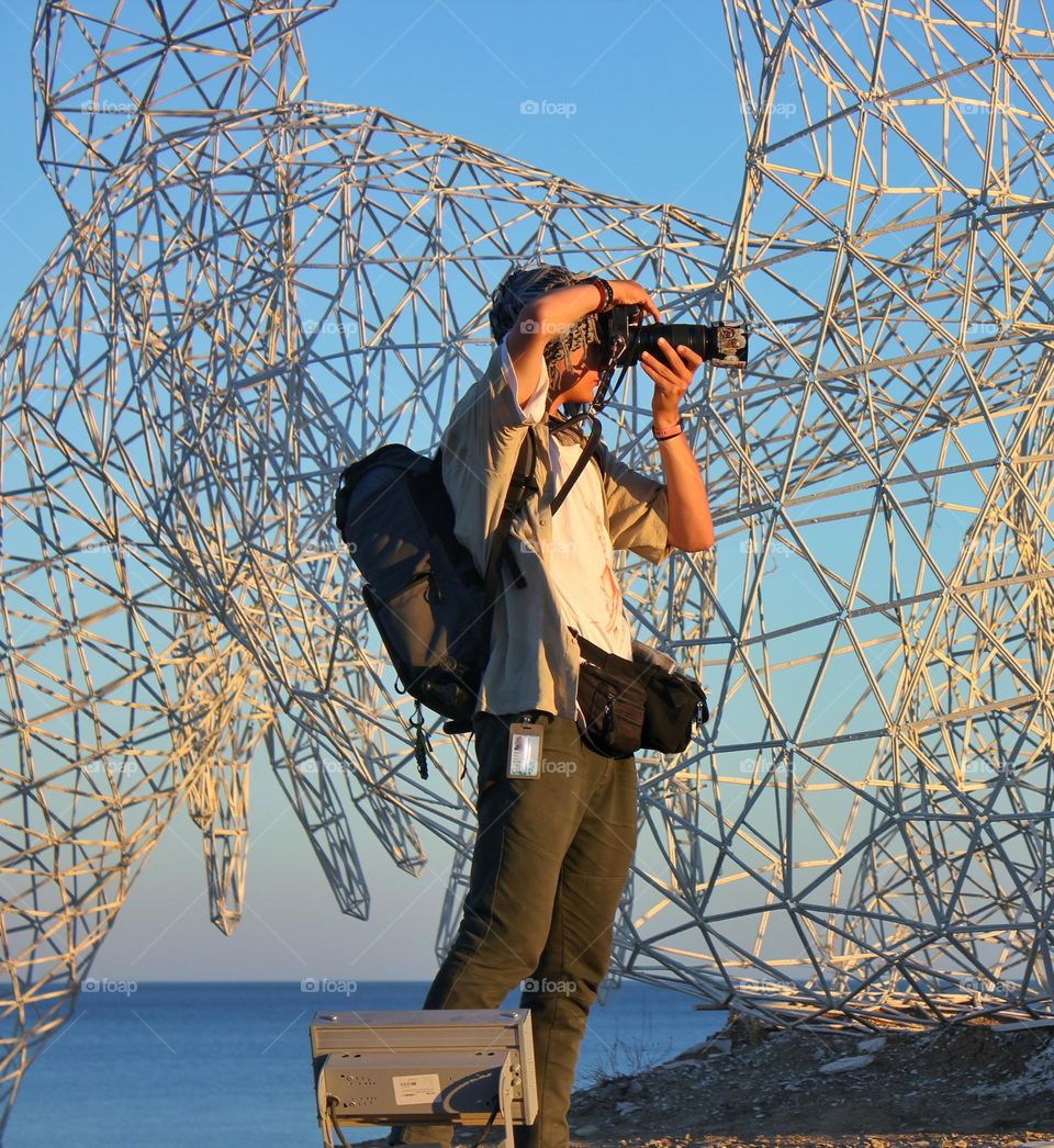 a photographer against the backdrop of a strange metal structure near the sea takes a photo at sunset