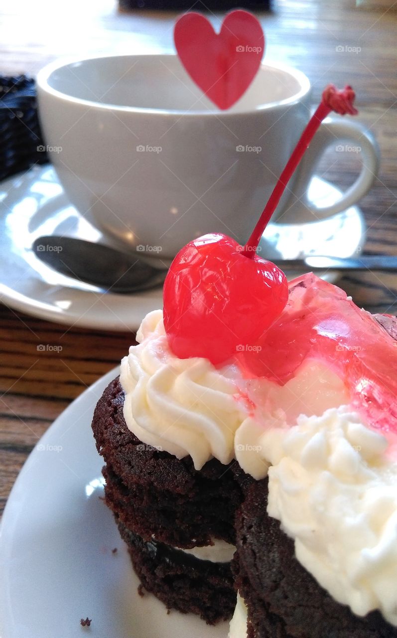 Chocolate cake with cherry on top. Cup of tea with heart on toothpick in the background.