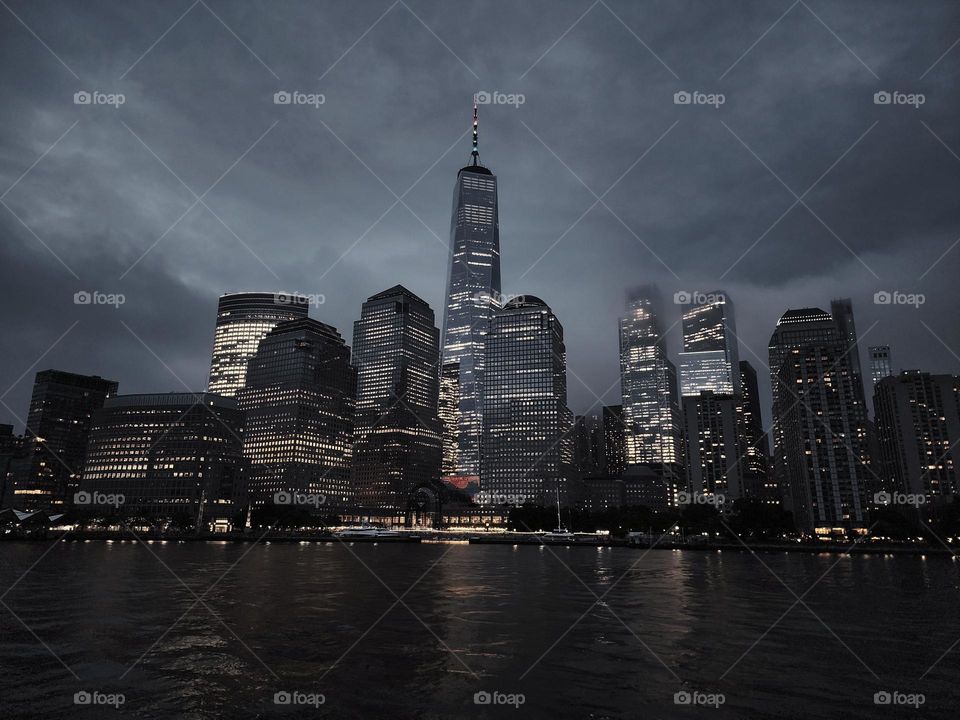 New York City skyline at night. Dramatic sky is the backdrop for the buildings, including the World Trade Center. The skyscrapers are illuminated by all the lights shining bright through the buildings many windows. The Hudson River in the foreground.