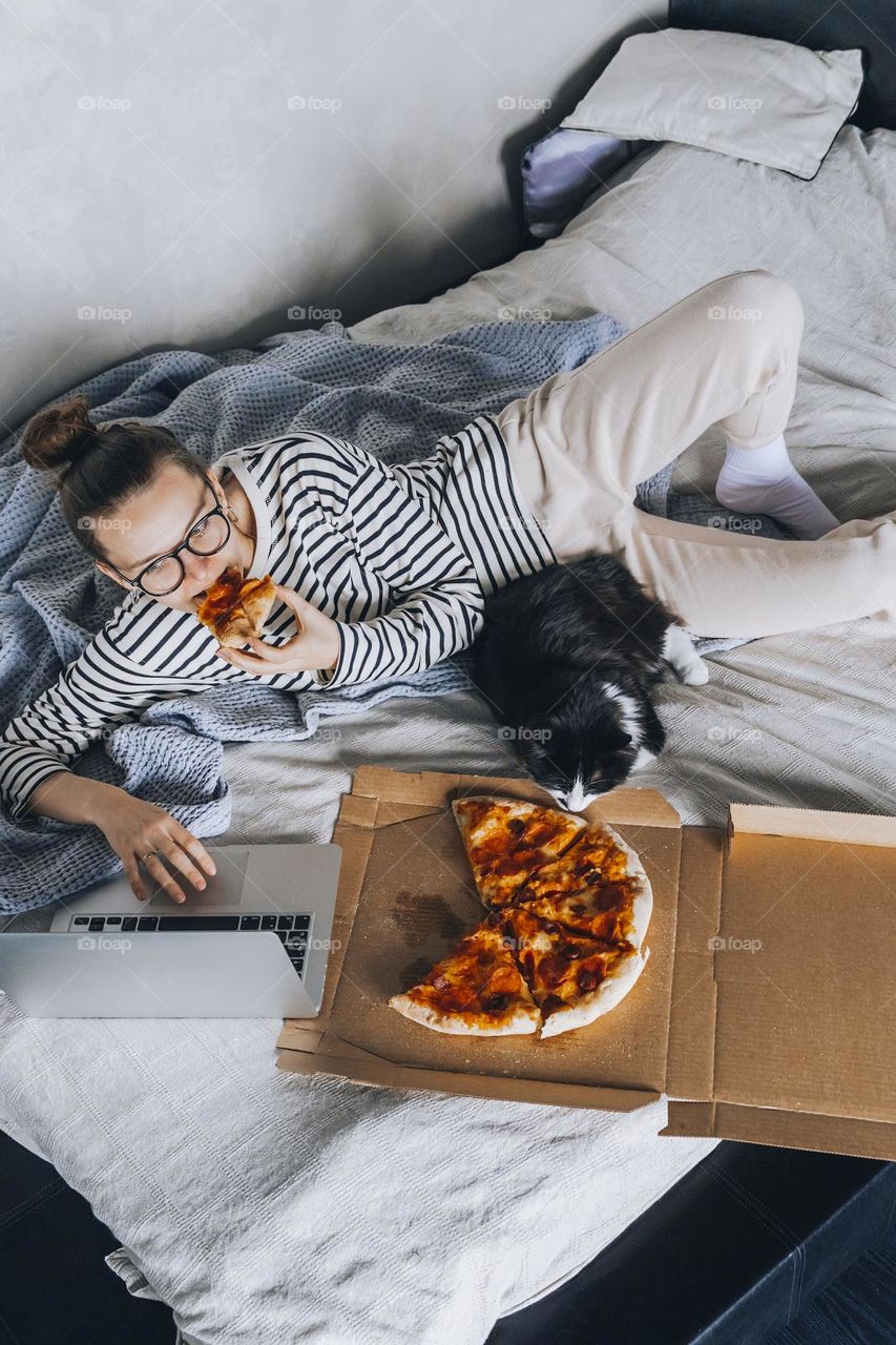 Millennial woman in bed eating pizza in bedroom. Apartment life 