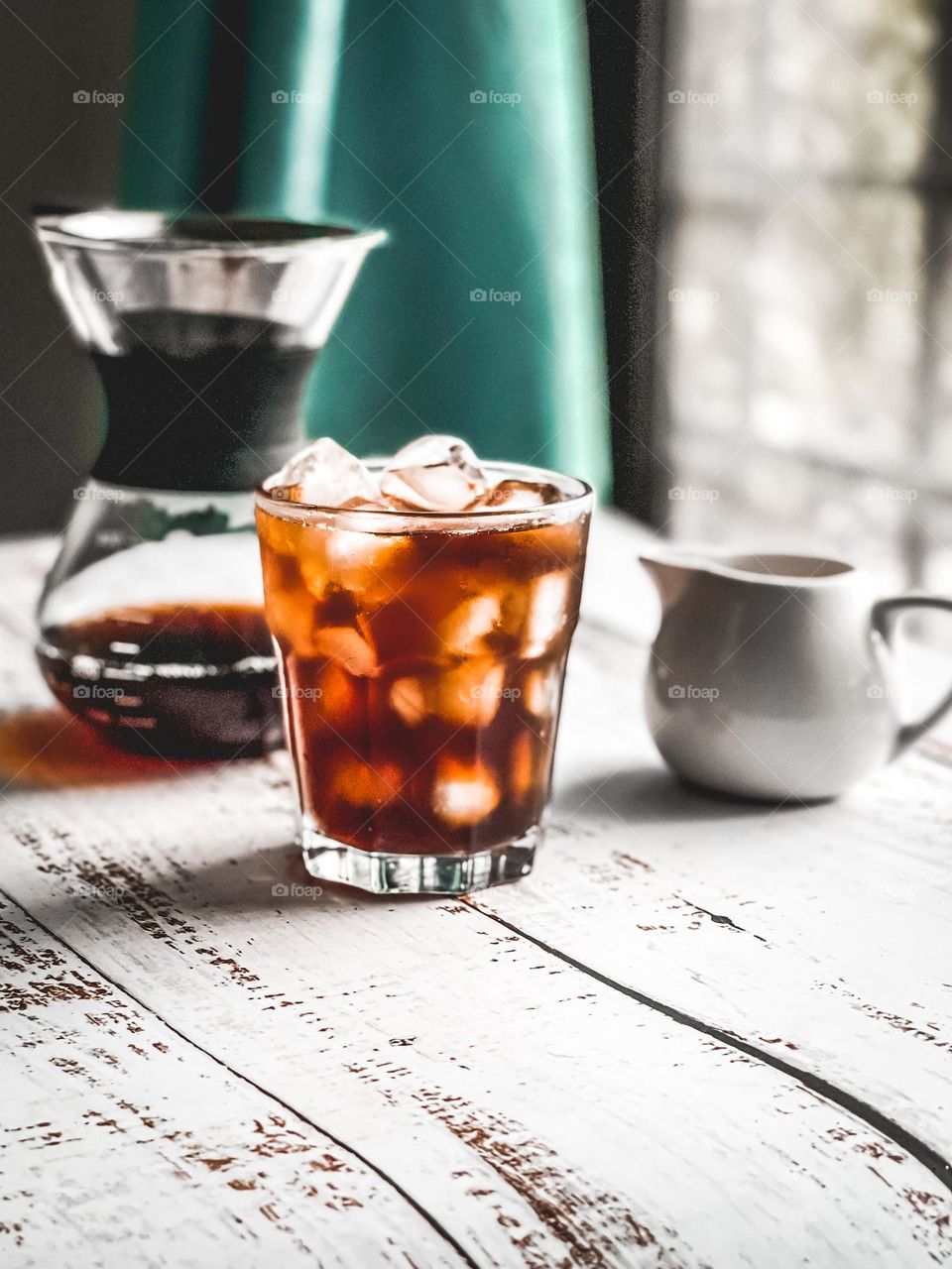 Iced Americano in a cup next to coffee dripper and a small milk jug on white wooden table with harsh light