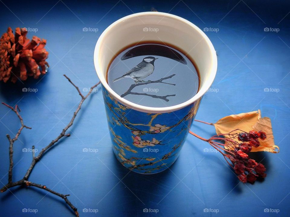 On a dark blue background with a reflective effect, there is a mug with a Japanese-style pattern. In a mug of black coffee on the surface of which is a reflection of a cherry branch and a bird.