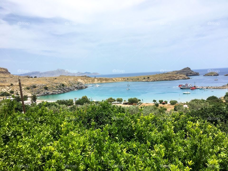 many green trees, purple-blue water surface, many yachts are anchored, there is a row of rocks, the sky is cloudy, blue-purple color , a beautiful day and the air is very fresh