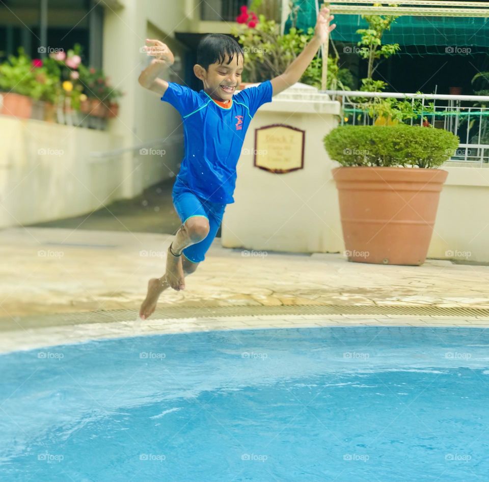 A 6 years old boy enjoying a lot at swimming pool in summertime and the background is filled with full of colourful flower pots and he jumping from high and smiles and wore blue colour swimsuit and he is very energetic and graceful.