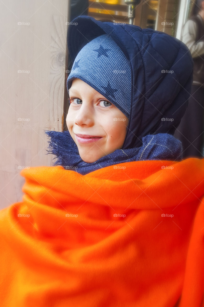 Portrait photo.  A boy in warm clothes with an orange throw blanket sits in the autumn in an outdoor cafe