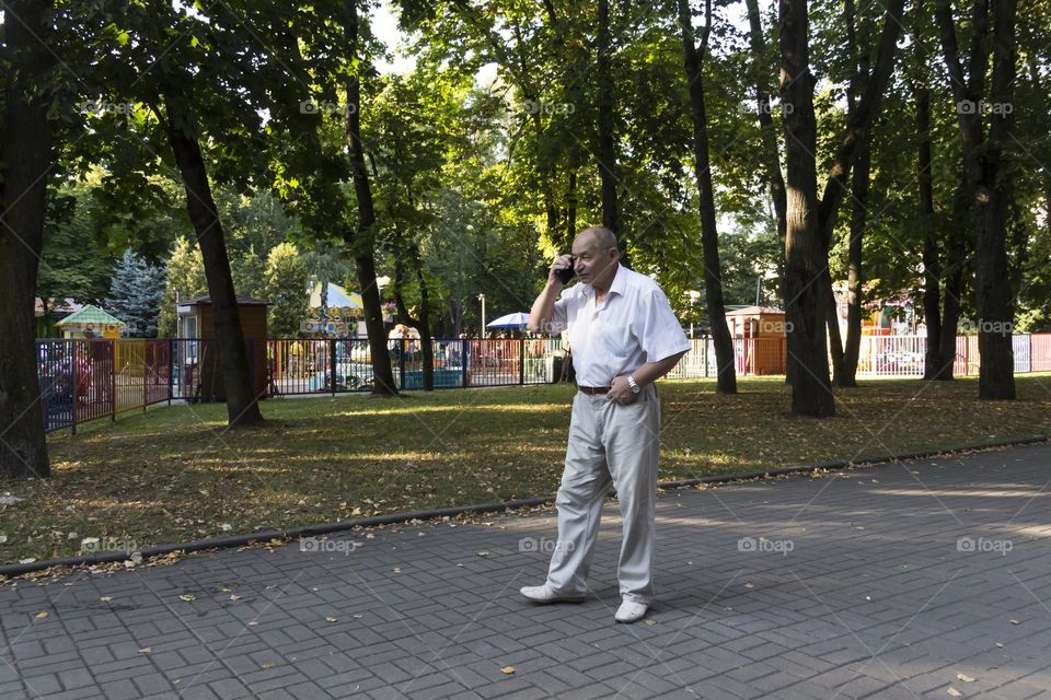 An elderly man walks alone in the park in the summer. A modern pensioner, businessman in a white shirt and trousers takes pictures with a camera in a mobile phone.