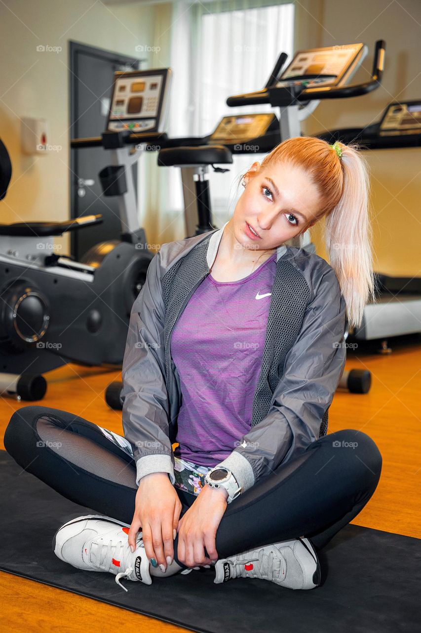 Blonde sports girl, with ponytail, sits on floor in fitness room, in front of fitness equipment