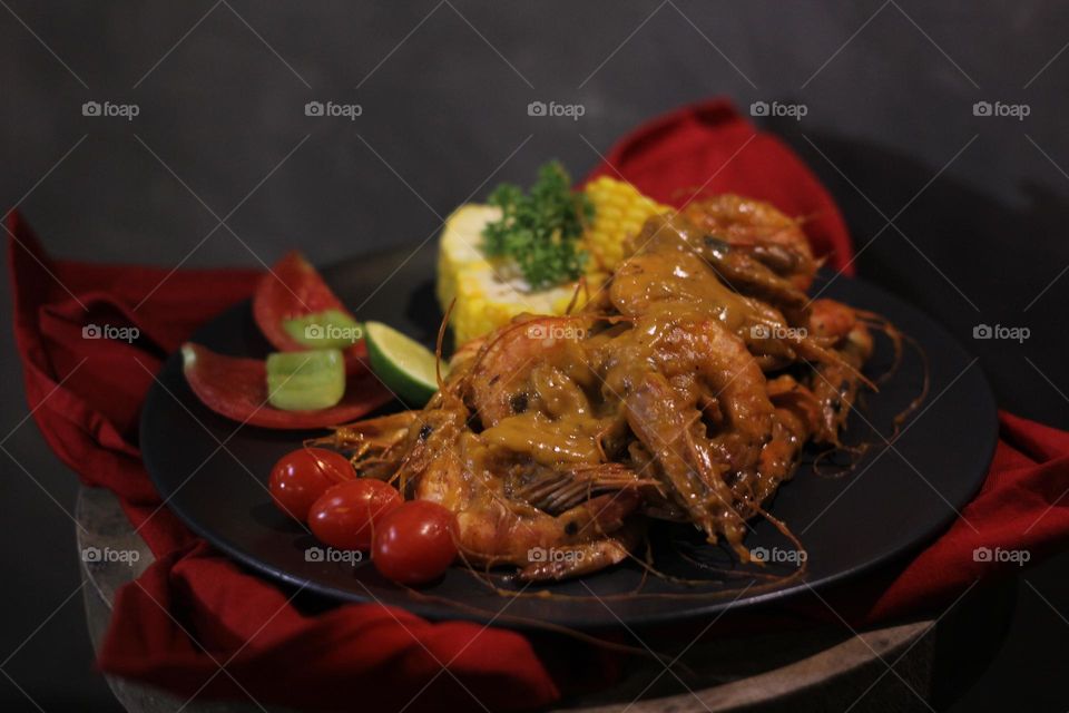 Asian food balado fried prawns with corn chunks, lemon slices and tomatoes served on black plate with red cloth bottom and wooden coaster. Asian style street seafood.